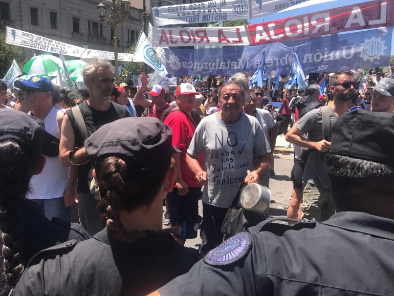 A man in a grey shirt bangs a pan while police look on at him and other protesters on the streets of Buenos Aires.