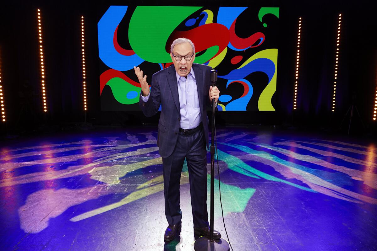 An angry-looking comedian in a dark blue suit poses with a stand-up microphone on a brightly colored stage.