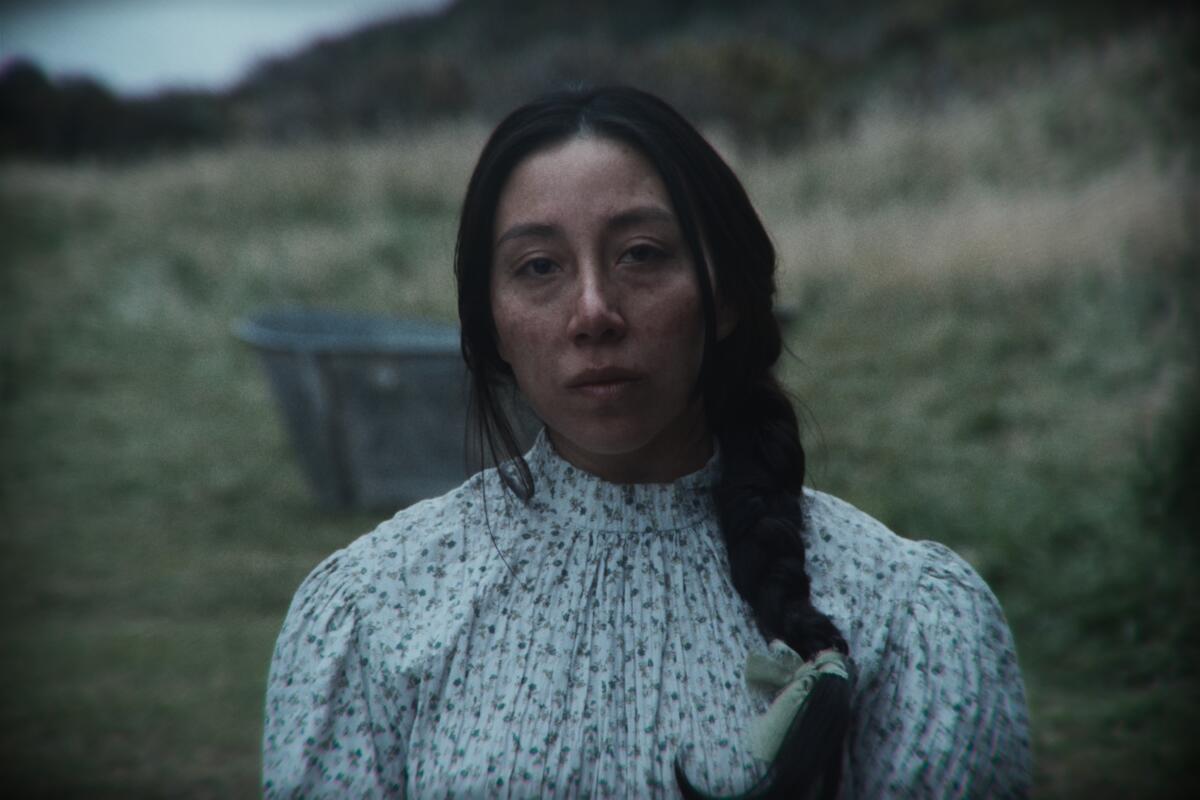 An Indigenous woman is seen in close-up in a floral dress against a grassy landscape.