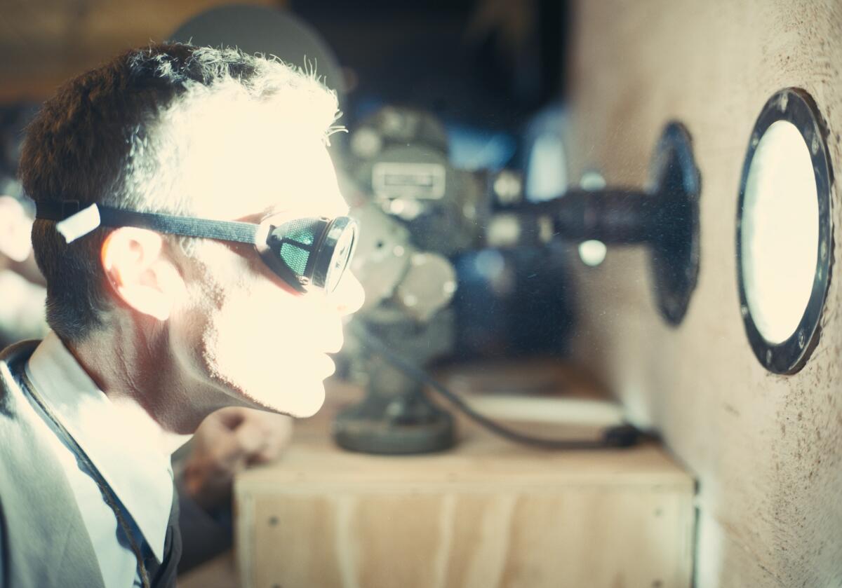 Cillian Murphy wears dark goggles as he peers through a porthole with intense light shining through in "Oppenheimer."