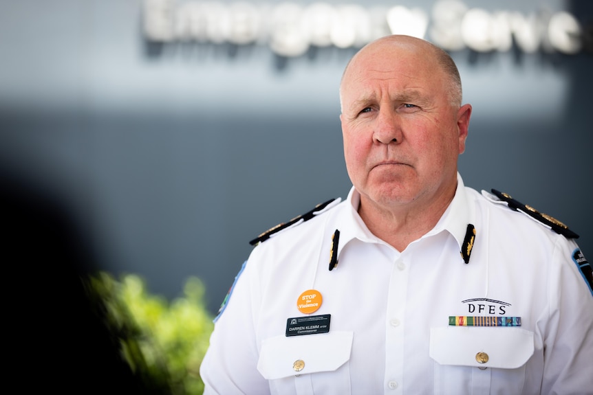 A man in a white shirt with badges and D-FES written on it with a stern expression while speaking.