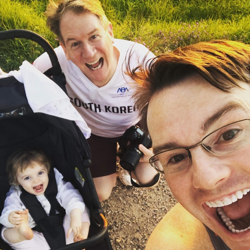 Two men smiling broadly as a toddler in a pram grins for a selfie.