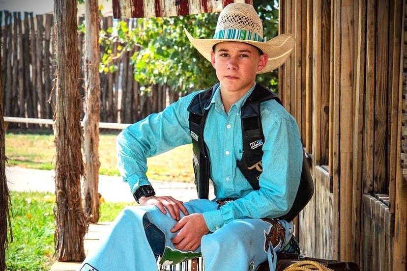 A young cowboy wearing a straw cowboy hat, blue shirt and jeans and brown vest.