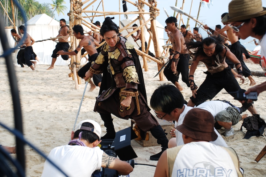 A man in armor with about a dozen other fights in the background, with film crew in the foreground.