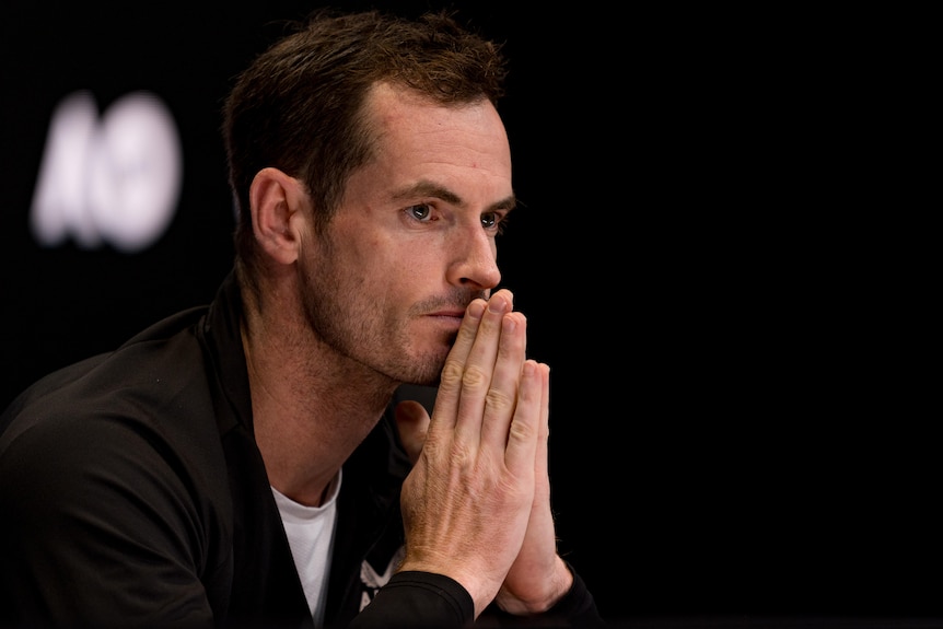 A man sitting at a desk, holding his hands together, during a press conference