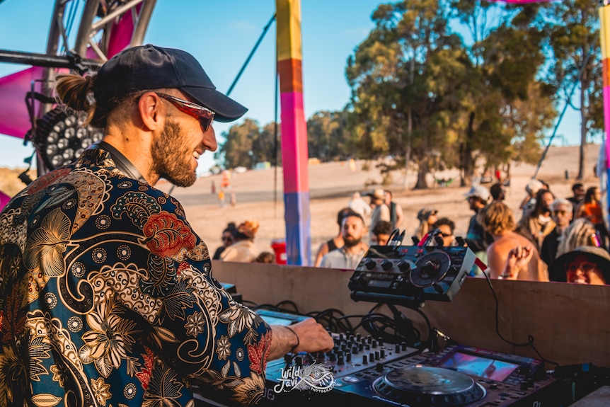 A DJ with hat and sunnies looks out at the crowd as he moves dials