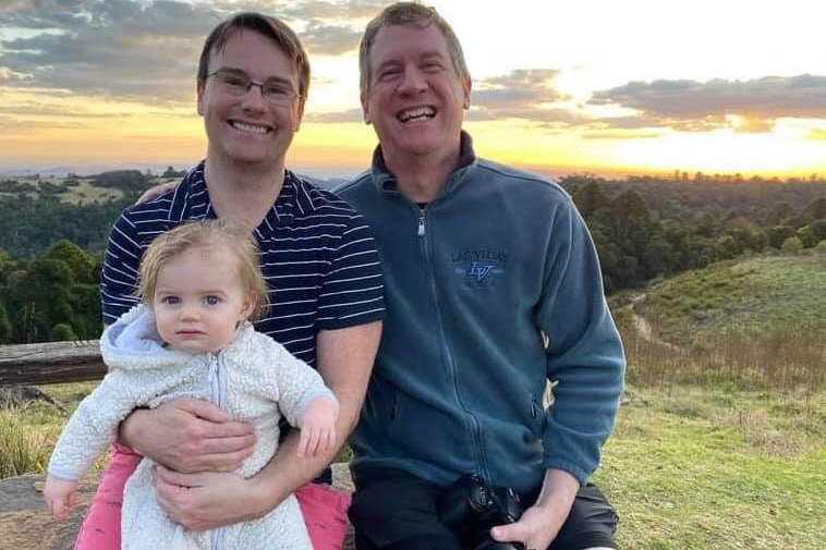 Two men hold a little girl as they sit ona  fence with a sunset behind them.