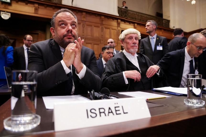 A man sitting at a desk, looking on, clasping his hands together, while other men sit near him.