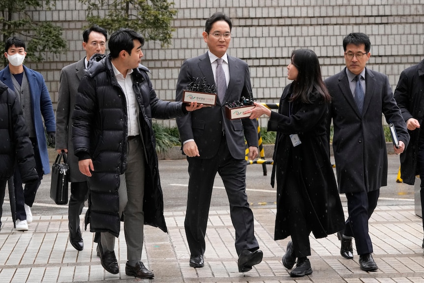 Four men in suits and one woman in a long coat walk on tiles. One woman and one man are holding a box with tiny microphones.