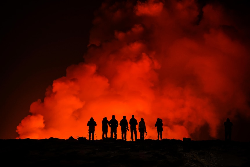 A black silhouette of seven people standing in front of a large plume of bright orange smoke