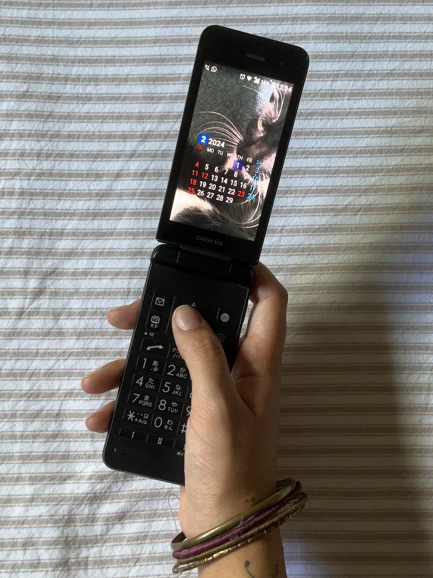 A close up of a hand holding and using a flip phone above the surface of striped bed sheets.