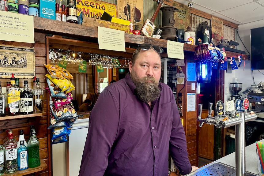 A bearded man leans on a bar.