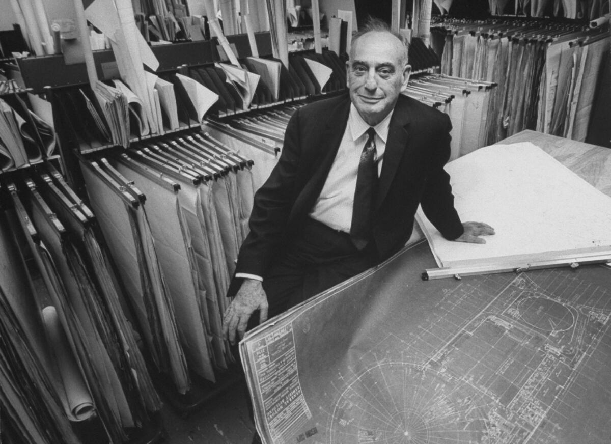 A black-and-white photo of a man in a dark suit and tie, seated near blueprints 