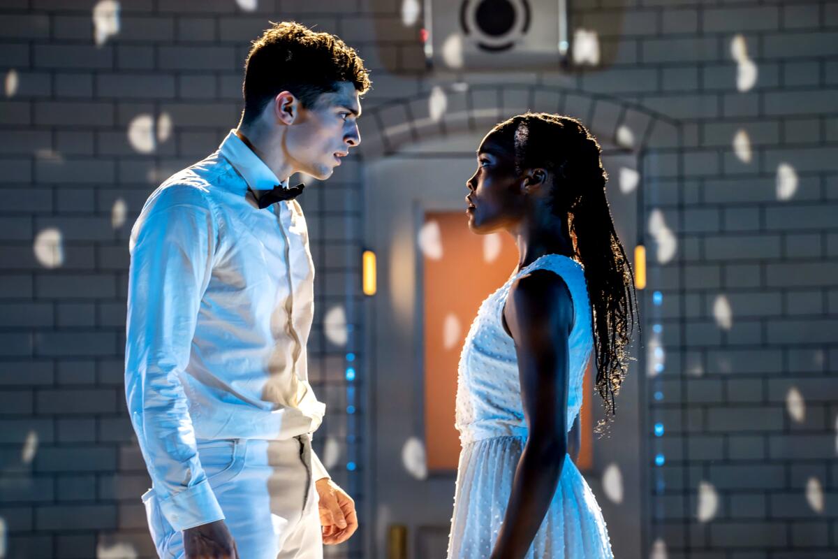 Two dancers — a white man and a Black woman — dressed in white, face each other on a dramatically lit stage.