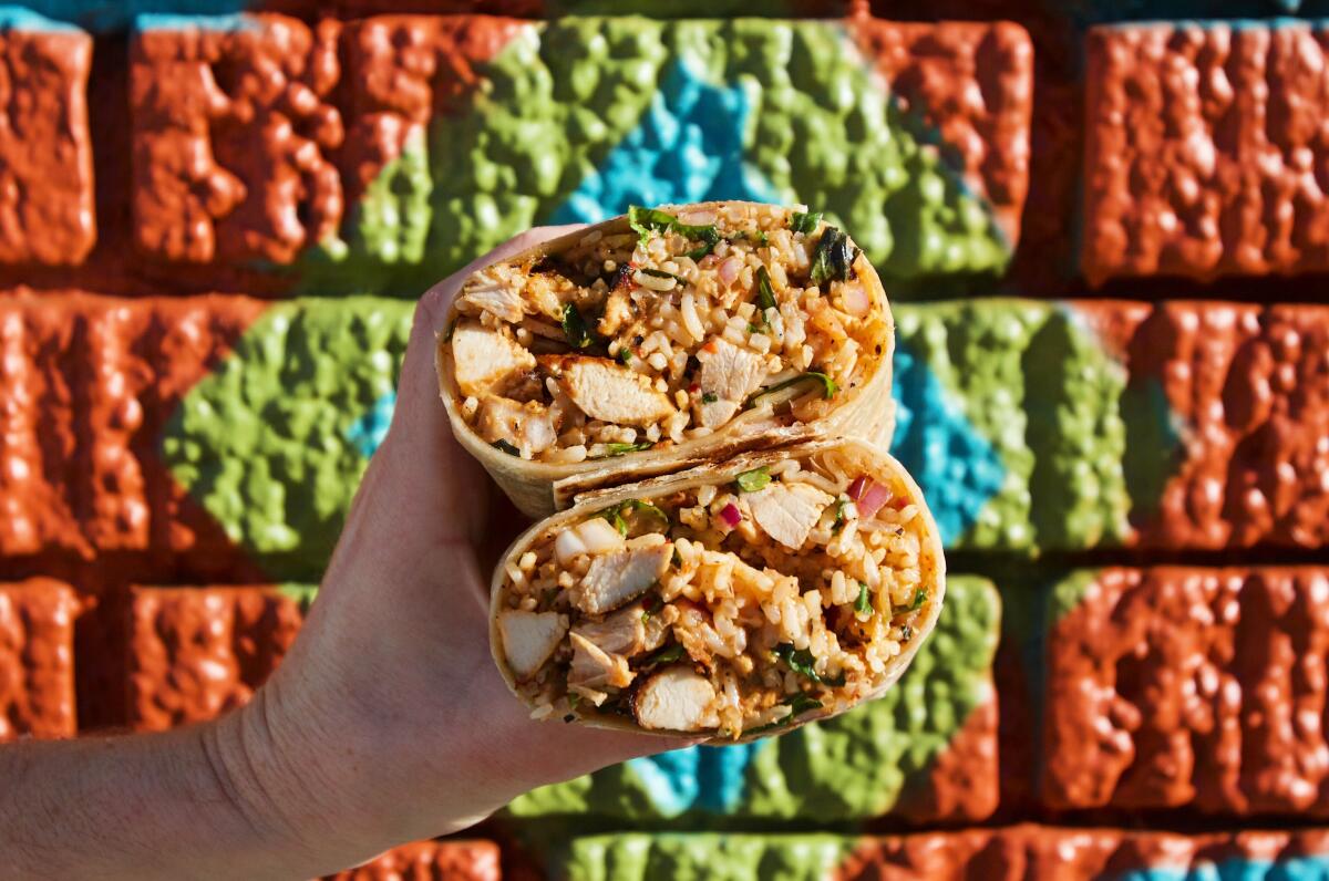 A horizontal pic of a hand with two stacked halves of a chicken burrito in front of a mural at Mid East Tacos in Silver Lake