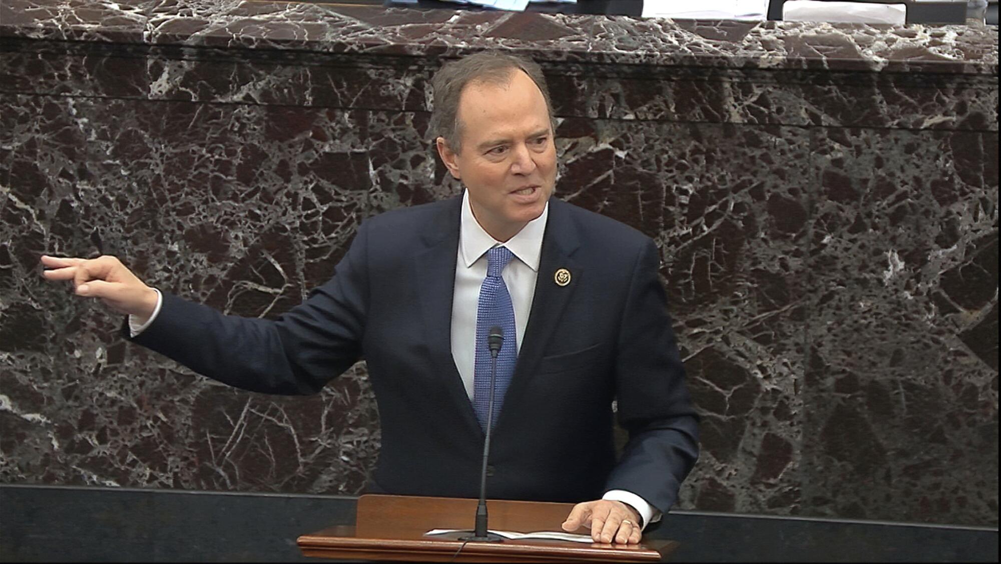  Adam Schiff gesturing in front of a dark marble wall as he speaks into a mic