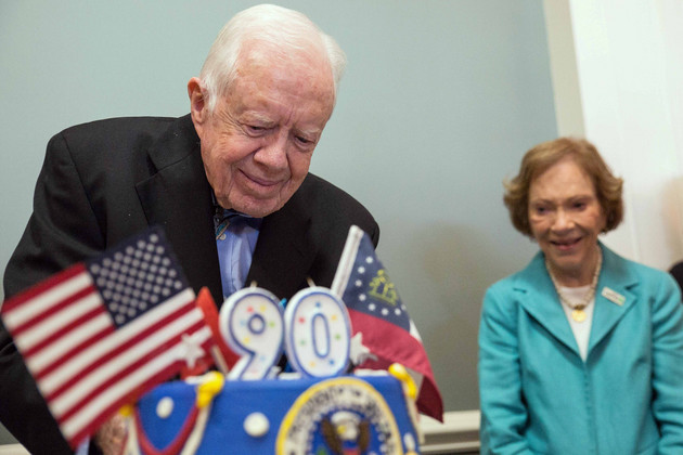 Jimmy Carter cutting a 90th birthday cake, with Rosalynn Carter standing in the background.