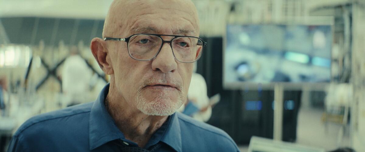 A man in glasses and a blue shirt stands in a room with screens behind him.