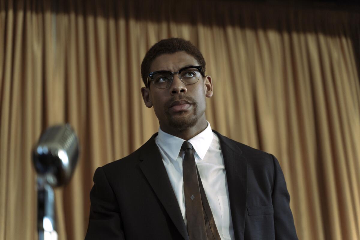 A man in a suit, tie and glasses standing before a microphone.