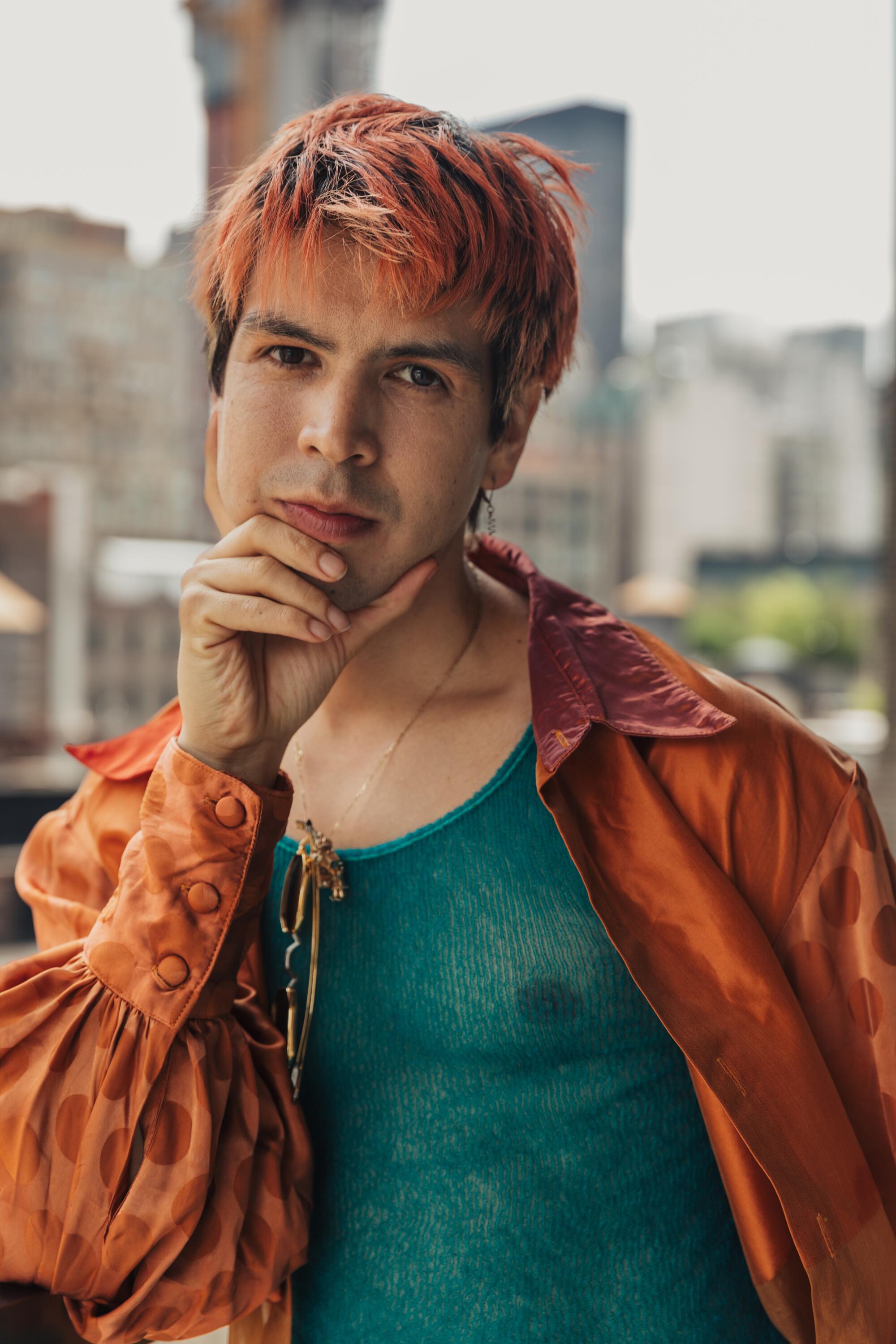 An actor-filmmaker in a green tank and silky orange shirt rests his chin in his hand.