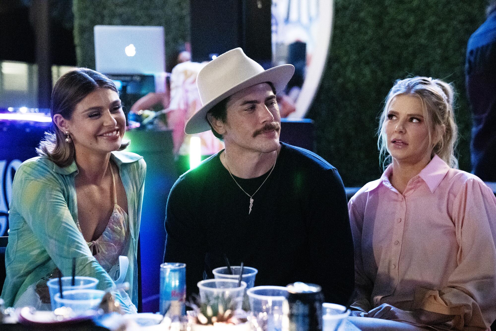 Raquel Leviss in a green shirt, smiling, seated next to Tom Sandoval in a black shirt and Ariana Madix in pink