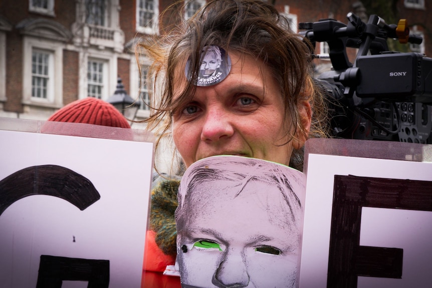 A woman with a sticker on her forehead and mask showing a man's face strung around her neck looks at the camera