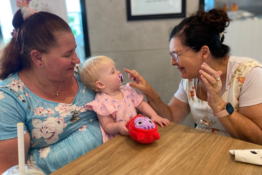 A mother and and her baby daughter sit at a cafe with another older woman playing with the child.