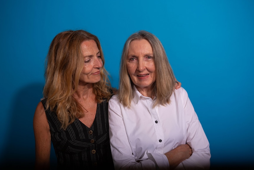 Against a blue background, Miranda Jakich, with shoulder-length grey hair and white shirt, smiles as woman next to her hugs her.