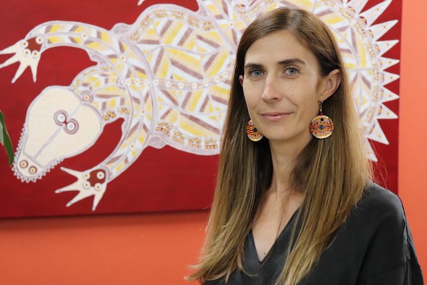 A woman with long hair tucked behind her ears looks at the camera in front of a wall painting 