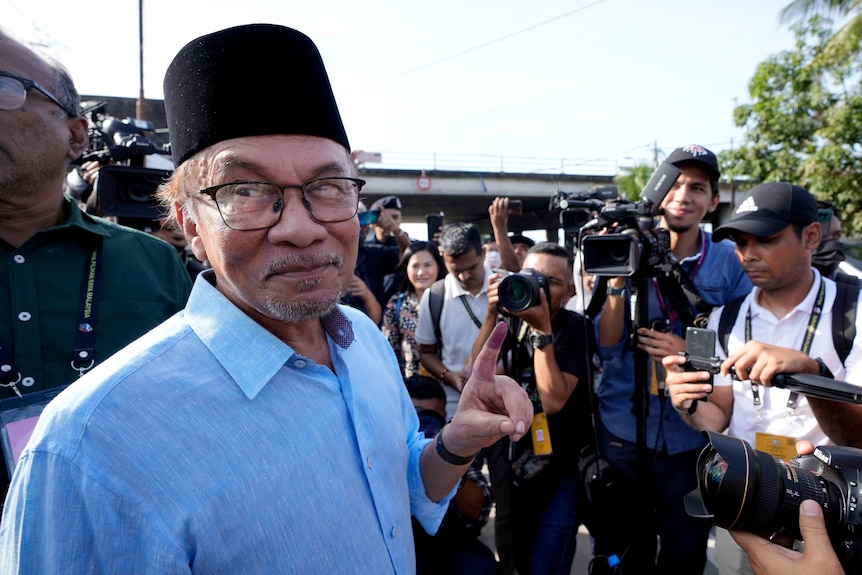 A man raises his left index finger while a press pack watches on.