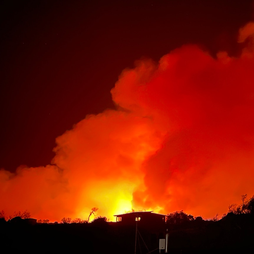 A house lit from behind by a huge, orange bushfire at night.