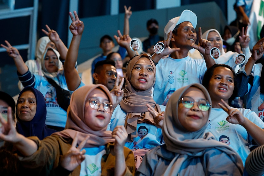 A crowd of people look at a screen displaying results and smile while waving their hands in the air.