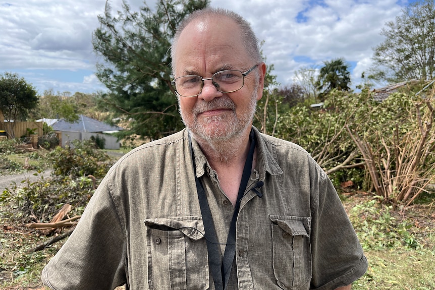 A slightly smiling bald man in glasses in front of damaged and fallen trees. He wears a crumpled khaki shirt.