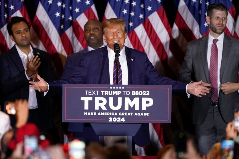 Donald Trump spreads his arms wide behind a podium emblazoned with his name. Behind him are political supporters and a line of US flags.