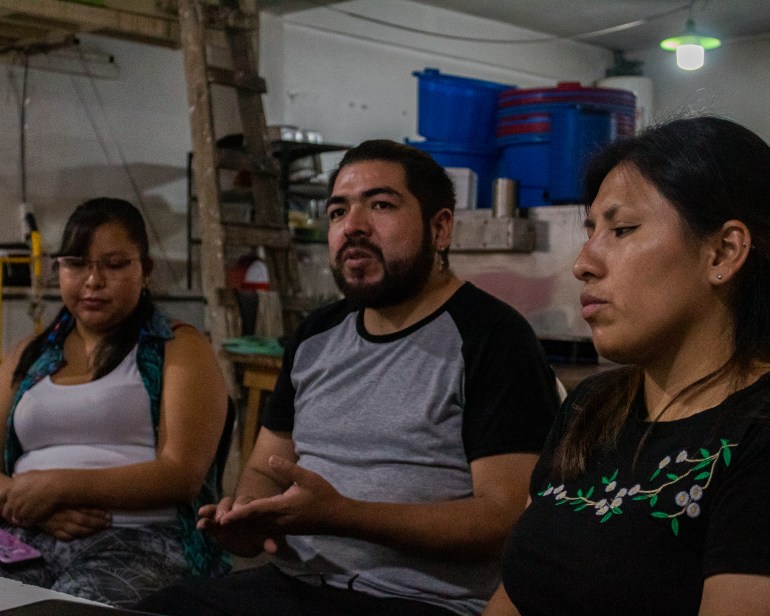 Three people sit in a darkened kitchen space: two women on either side of a man who is speaking.