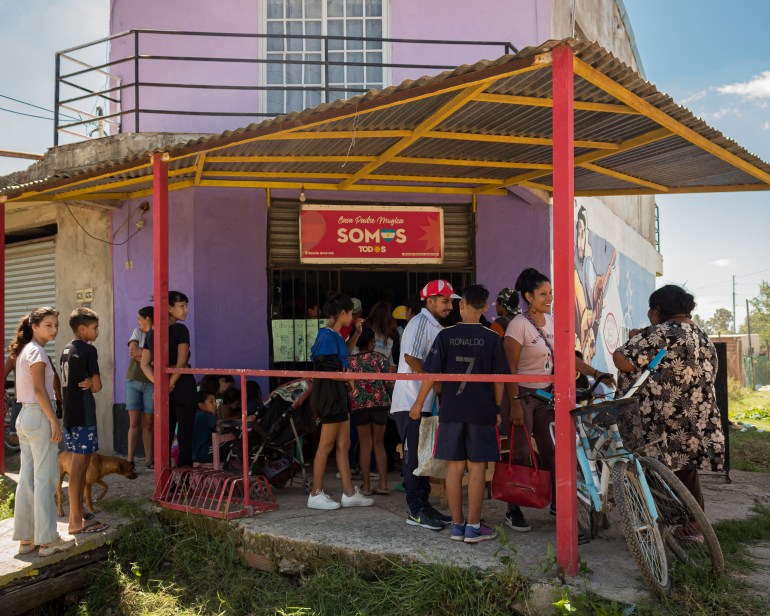 A queue of people wait outside a soup kitchen, located in a purple, multi-story building with an awning.