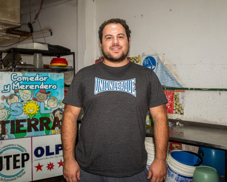 A man in a grey shirt stands in a cluttered room.