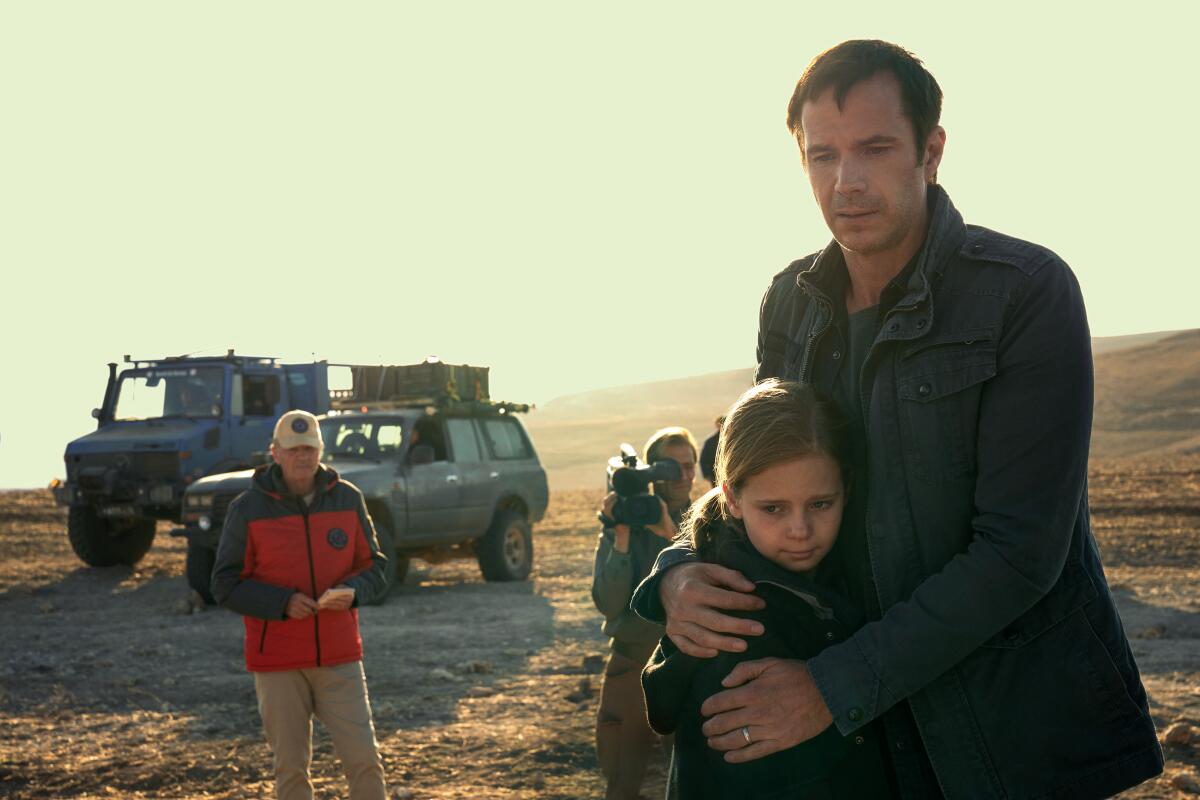 A man embraces a young girl as they walk through a desert with two men looking on behind them.