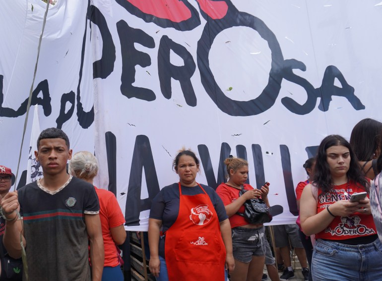 A woman in a red apron stands in front of a hand-painted protest banner.