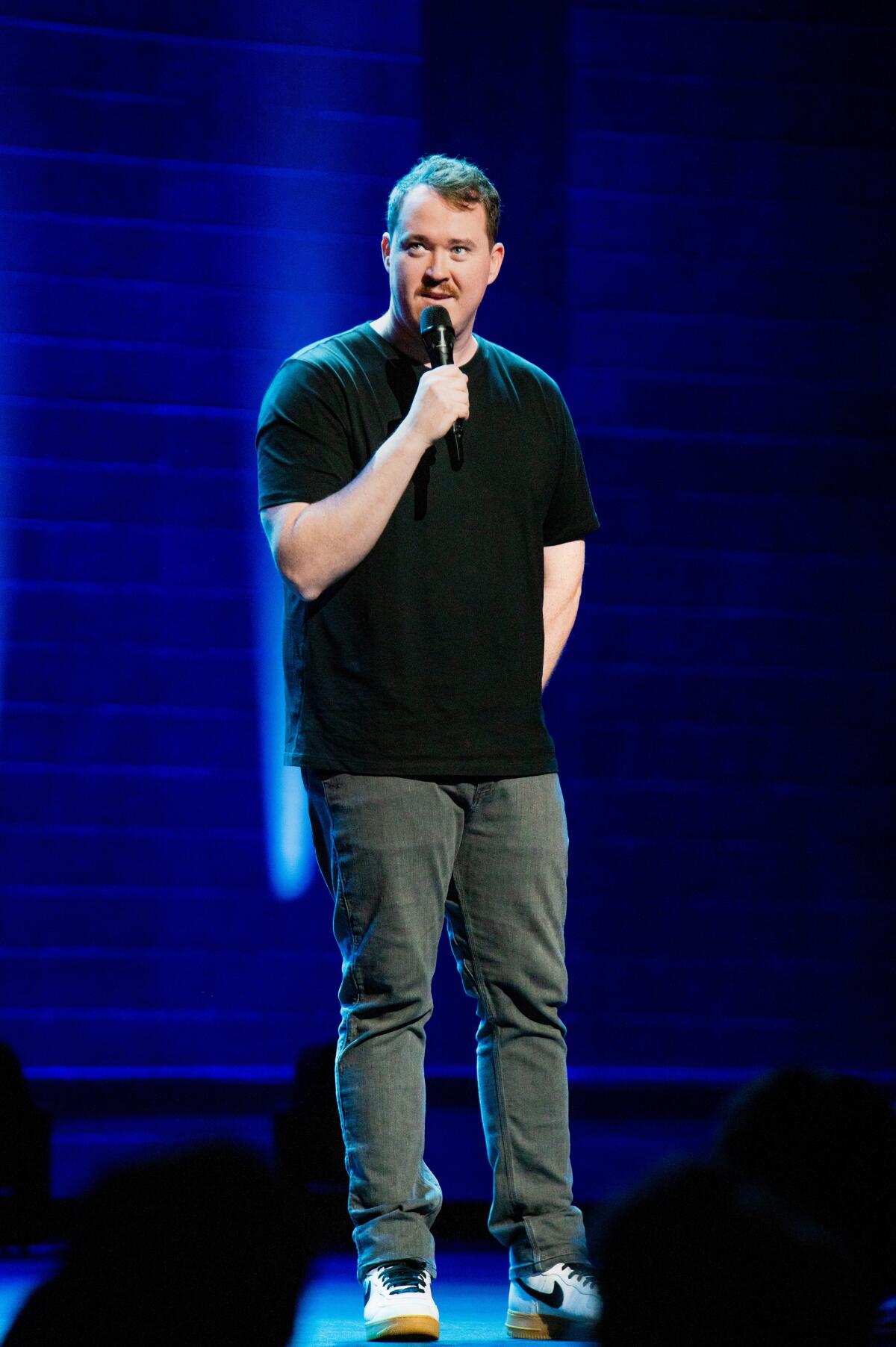 A man holding a mic stands on a stage in a black T-shirt, jeans and sneakers.