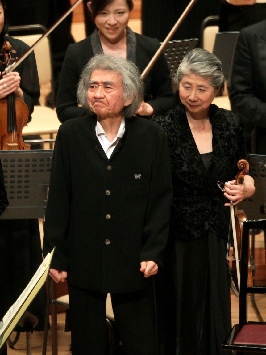 An old man pursing his lips to hold back tears as he is surrounded by people holding instruments and clapping