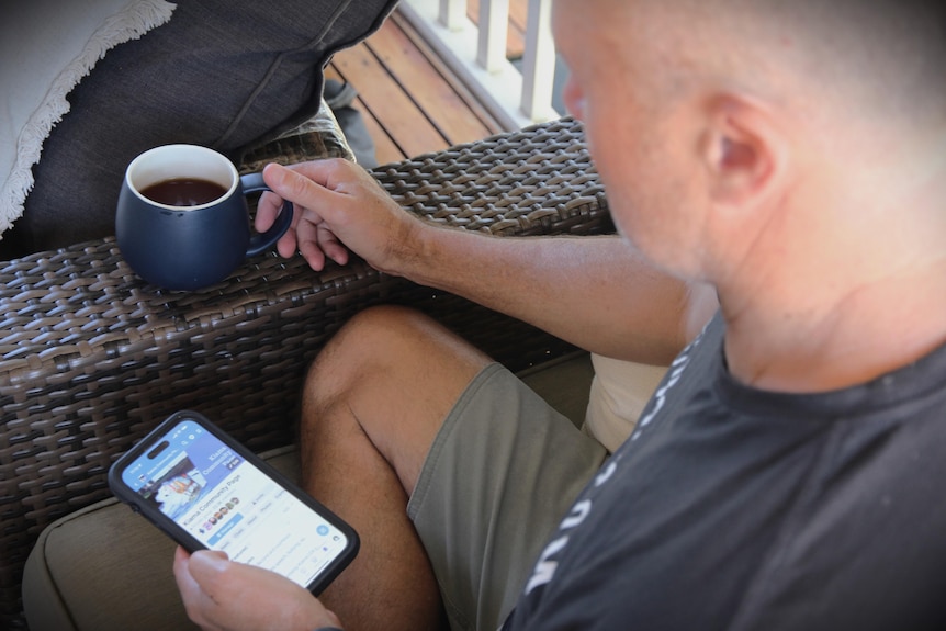 Man holding cup of coffee in right hand, sitting on chair, holding Facebook open on phone in left hand