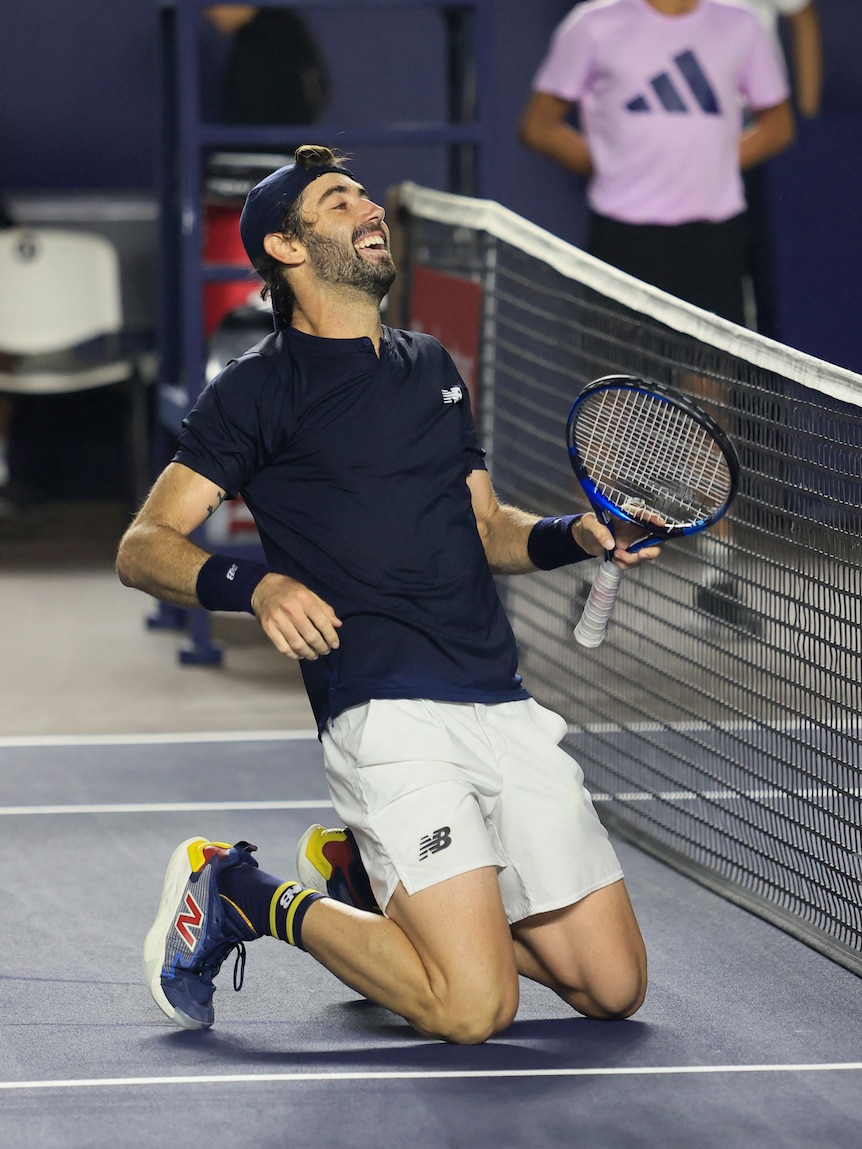 Jordan Thompson, dark shirt and white shorts, on his knees smiling in celebration at the net.