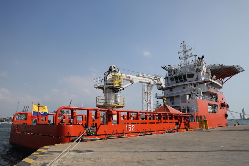 A large orange ship with a yellow, red and blue flag floating on it.