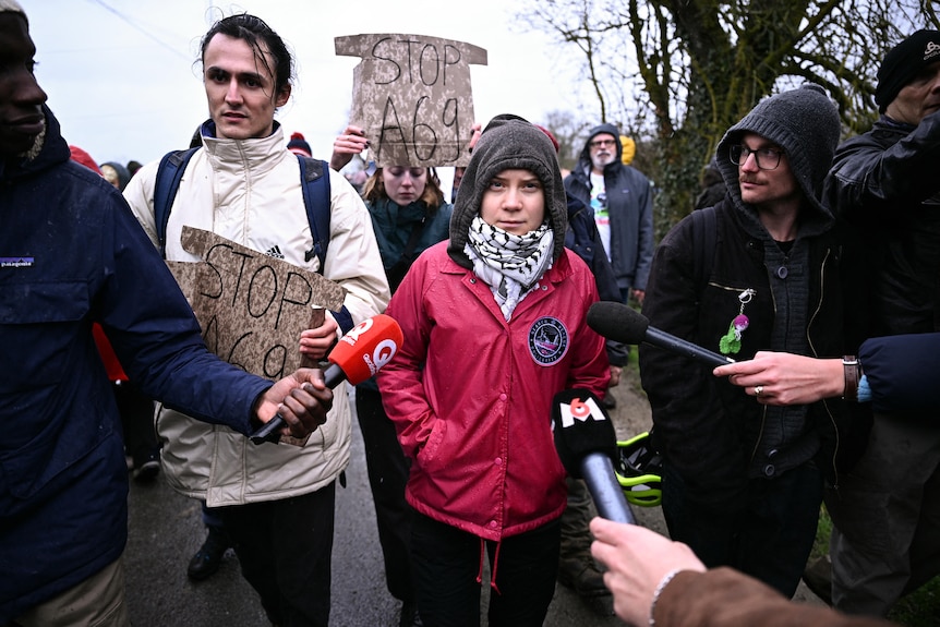 A young woman wearing a red jacket and black and white scarf surrounded by a crowd of people carrying signs