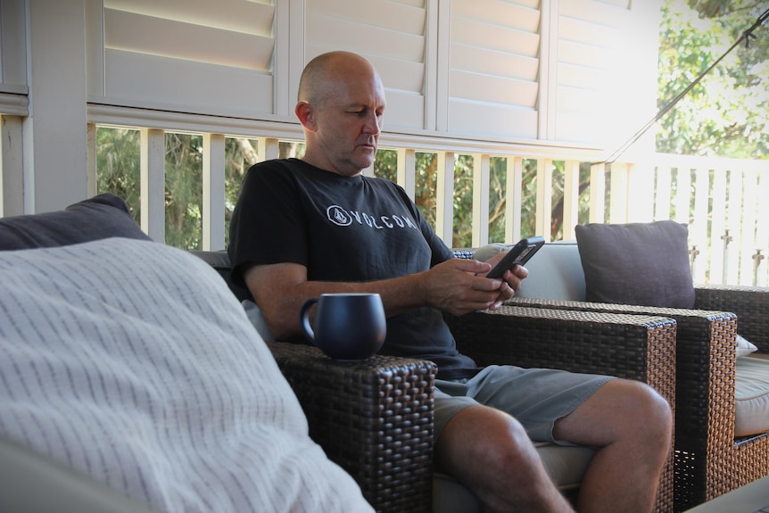 Bald man wearing black shirt sitting down on verandah holding phone