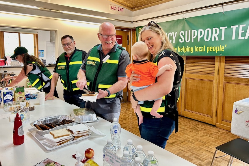 A woman holding a baby talks to a man who is preparing food.