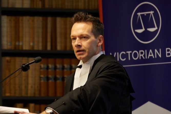 A man with short dark hear wearing a black suit speaks at a podium with a bookshelf in background