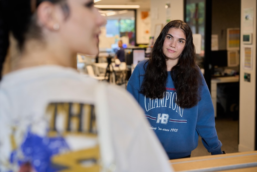 A slightly young woman in a blue sweater looks towards another woman wearing a white hoodie in a common kitchen area.