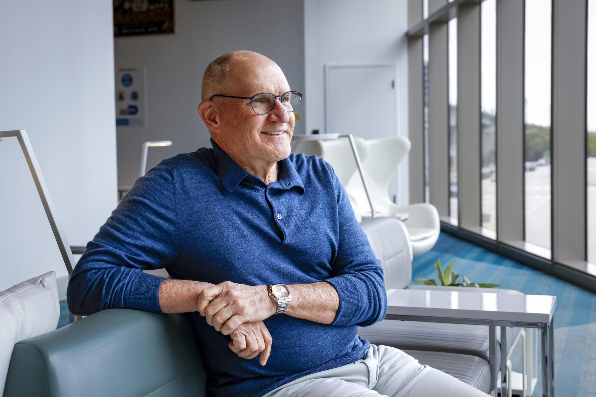 A man in a blue long-sleeved shirt and glasses, hands clasped and smiling, is seated facing windows